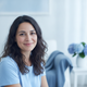 A smiling French woman in her late 20s, wearing a light blue top, with a blurred soft blue and white background.